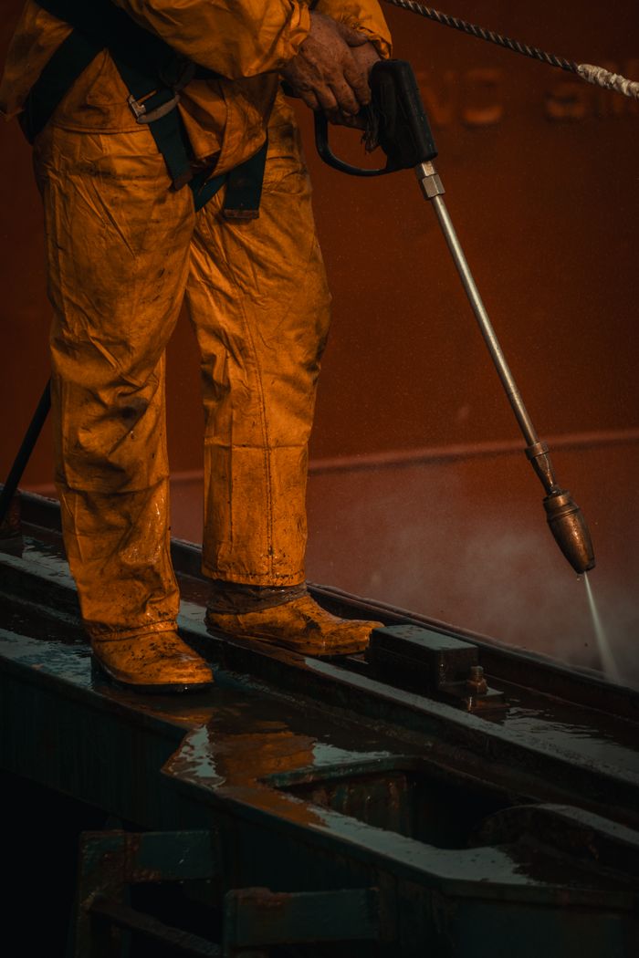gallery-01 A worker in yellow safety gear uses a pressure washer on industrial equipment.